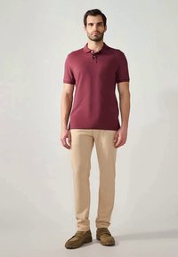 Uomo che indossa una polo bordeaux, pantaloni beige e mocassini marroni, in piedi contro uno sfondo chiaro e uniforme.