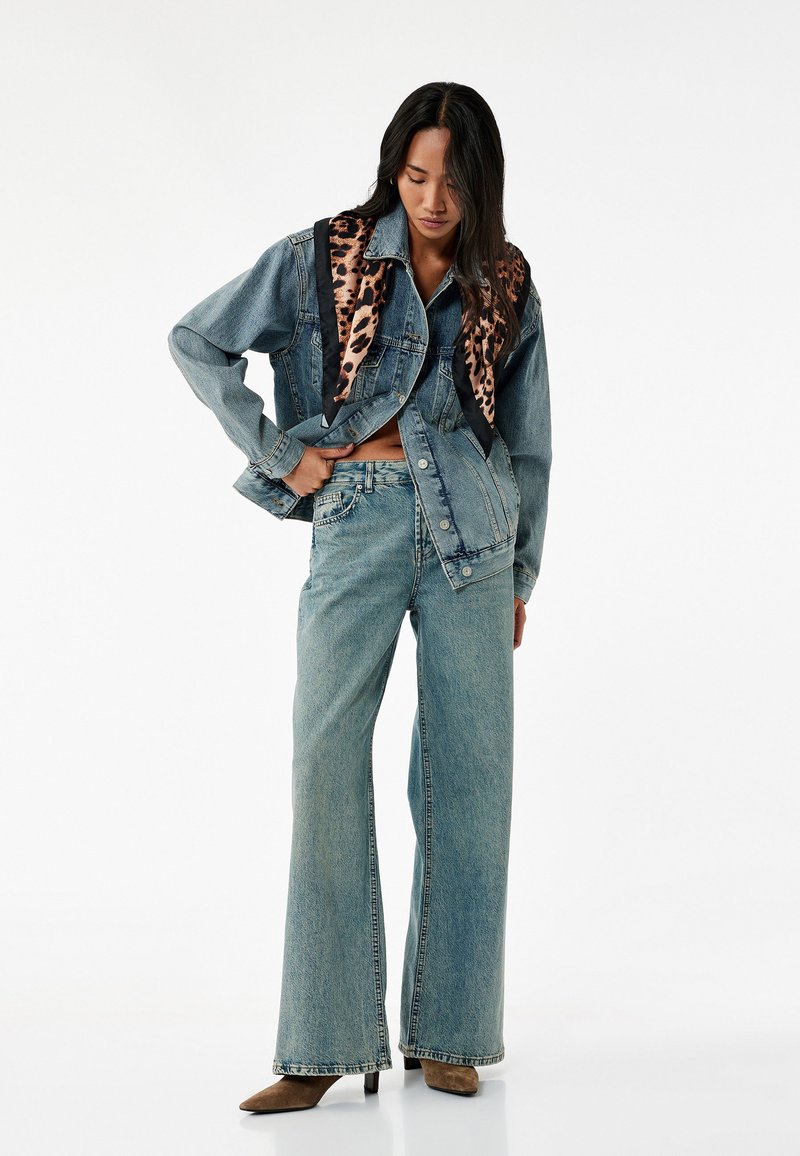 Farkkutakki leopardikuvioisilla yksityiskohdilla, löysä istuvuus, vaaleansiniset farkut ja ruskeat korkokengät, korostaen erottuvia tekstuureja ja kuvioita.