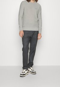 Man i ljusgrå långärmad tröja, mörkgrå byxor och vita och svarta sneakers som står framför en enfärgad bakgrund.