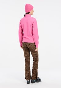 Fleece pullover, kirkas vaaleanpunainen, korkea kaulus. Leopardikuosiset housut ruskeassa ja mustassa, nilkassa vetoketjut, yhdistettynä neulottuun vaaleanpunaiseen hattuun.