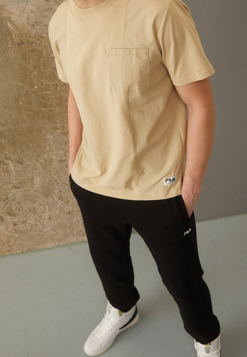 T-shirt en coton beige avec une poche poitrine et un pantalon de survêtement noir avec un logo brodé. Le modèle porte des baskets blanches.
