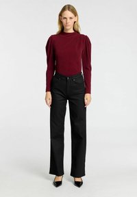 Burgundy långärmad topp med puffade axlar, kombinerad med högmidjade svarta jeans och svarta spetsiga klackar, mot en neutral bakgrund.
