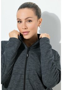 Giacca grigia con zip realizzata in un tessuto texturizzato. Presenta un colletto alto, maniche aderenti e una chiusura frontale con zip. La modella tiene il colletto vicino al viso.