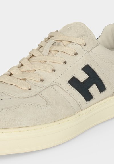 Gros plan d'une basket en daim beige avec des lacets crème, logo noir "H" sur le côté et semelle en caoutchouc blanc cassé.