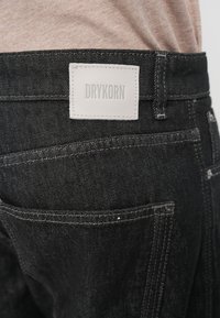 Jeans in denim nero con un'etichetta in pelle bianca sul retro con la scritta "DRYKORN". Presentano cuciture visibili e un classico design a cinque tasche.