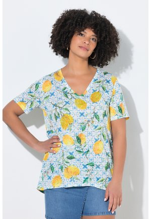 Frau mit lockigem Haar, die ein V-Ausschnitt-Shirt mit gelben Zitronen und blauem Blumenmuster sowie einem blauen Jeansrock trägt, posiert mit der Hand in der Hüfte.