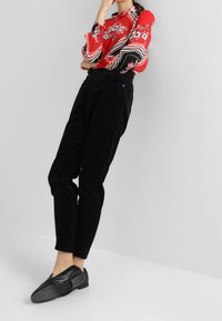 Blusa floreale rossa con righe bianche e nere; abbinata a pantaloni in corduroy neri a vita alta; modella che indossa scarpe nere flat, braccia incrociate.