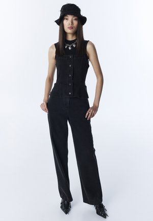 Młoda kobieta ubrana w czarny bezrękawnik zapinany na guziki, szerokie czarne spodnie, czarne buty na szpic, czarny kapelusz typu bucket oraz srebrny naszyjnik.