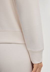 Manche de sweat-shirt beige clair avec poignet, fabriquée en tissu doux, texture lisse et détails de couture subtils, sur fond uni.
