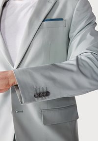 Jachetă de costum gri deschis cu o textură netedă. Include o batista albastră în buzunar, patru nasturi întunecați pe manșetă și un singur buzunar frontal.