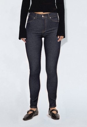 Kvinde iført mørkeblå skinny jeans, sort cropped langærmet top og sorte flade sko med spænder, stående mod en ensfarvet baggrund.