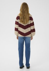 Osoba z długimi blond w�łosami, ubraną w sweter w beżowo-bordowe zygzaki, niebieskie dżinsy i czarne buty, stoi tyłem.