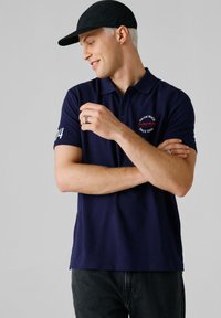 Marineblaues Poloshirt mit kurzen Ärmeln, mit einem gestickten Motiv auf der Brust und einer weißen Zahl auf dem Ärmel. Der Stoff wirkt strukturiert.