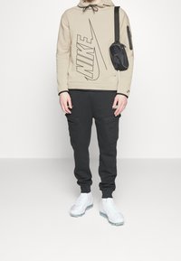 Beige hoodie med svart Nike-logga, svarta cargo-byxor och vita sneakers. Accessory-väska fäst vid axeln. Varm, avslappnad outfit.