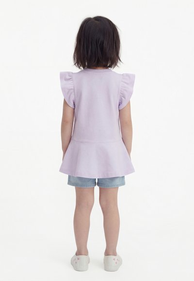 Παιδί με κοντά σκούρα μαλλιά, φορώντας ένα ανοιχτό μωβ τοπ με βολάν στα μανίκια, τζιν σορτς και άσπρα παπούτσια με ροζ σχέδια καρδιάς, με την πλάτη προς τα εμάς.