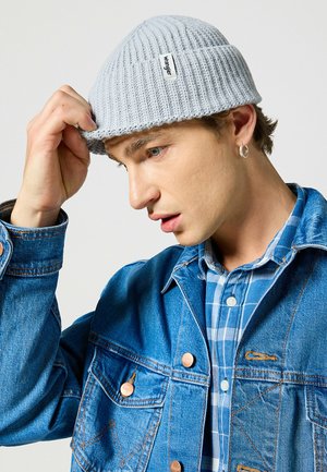 Berretto lavorato a maglia di colore grigio chiaro con texture a coste. Il modello regola il berretto mentre indossa una giacca di jeans sopra una camicia a quadri blu.