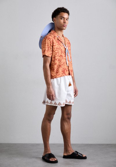 Short-sleeve orange shirt with floral pattern, paired with white shorts featuring brown embroidery, black sandals and a light blue hat.