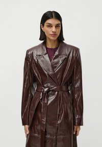 Una donna con capelli lunghi e lisci di colore scuro, che indossa un cappotto in pelle lucida di colore marrone scuro con cintura sopra un top viola, su uno sfondo neutro.