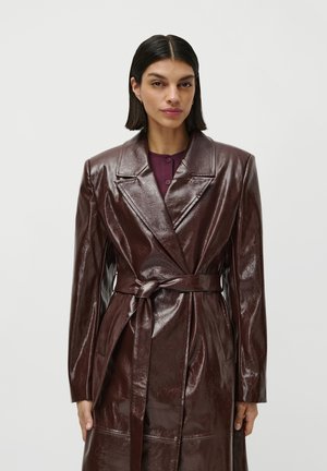 Una donna con capelli lunghi e lisci di colore scuro, che indossa un cappotto in pelle lucida di colore marrone scuro con cintura sopra un top viola, su uno sfondo neutro.