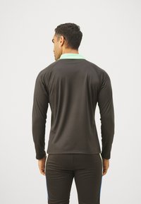 Langermet treningsskjorte i svart med lys grønn krage. Har en glatt tekstur og en tettsittende design. Bakre utsikt av skjorten.