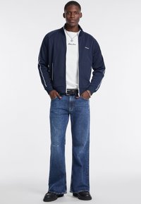 Jūras spēku zip-up jakas ar baltām akcentiem, valkāta virs balta t-krekla. Apvienota ar brīvām zilu džinsu biksēm un melnām kurpēm, kas raksturojas ar gludu tekstūru.