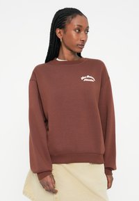 Sweatshirt castanho feito de tecido suave, com decote redondo, mangas largas e um logotipo bordado branco no peito.