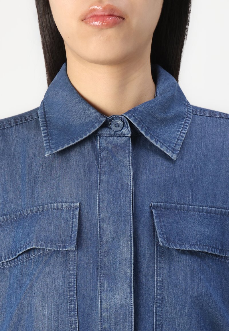 Chemise en denim avec un col à boutons, deux poches poitrine et une surface lisse et texturée dans une teinte bleu moyen.