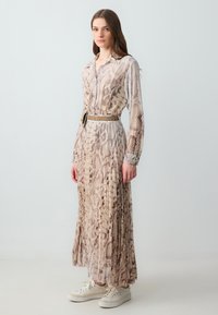 Lungo vestito beige con un motivo a stampa di serpente. Progettato con una chiusura a bottoni sul davanti, maniche lunghe e una vita con cintura. Abbinato a sneakers bianche.