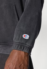Dunkelgrauer Sweatshirtärmel mit geripptem Bündchen und einem kleinen gestickten Logo mit blauen und roten Elementen. Glattes Baumwollmischmaterial.