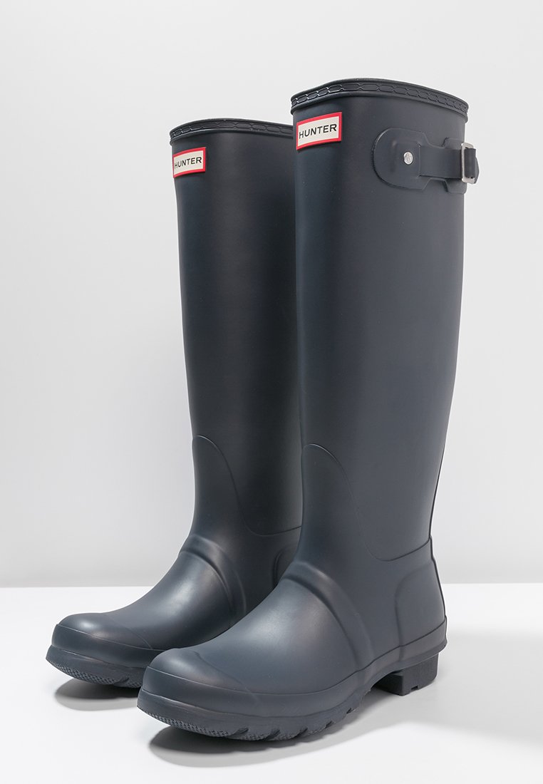 Botas de lluvia de goma negras con una textura suave, de altura media hasta la pantorrilla y un detalle de hebilla en el lateral. Presenta una etiqueta de marca distintiva.