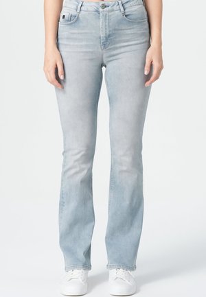 Hellblaue Flared Jeans, getragen von einer Person, die mit entspannten Händen an den Seiten steht, kombiniert mit weißen Sneakers auf schlichtem Hintergrund.