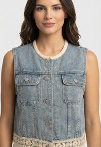 Mujer con chaleco vaquero sin mangas con botones, bolsillos con solapa y ribete de crochet crema en el escote y el dobladillo, sobre un fondo claro y liso.