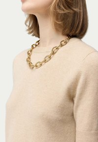 Guldkedja med stora ovala länkar som bärs över en beige tröja. Har en slät, reflekterande yta och en kraftig design.