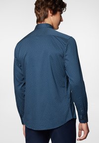 Granatowa koszula z długim rękawem w białe kropki, z kołnierzykiem odwróconym w dół i rozcięciami po bokach, wykonana z gładkiego materiału o delikatnym połysku.