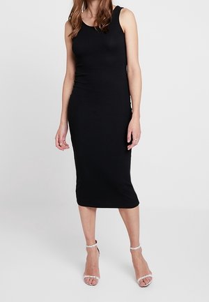 Frau trägt ein ärmelloses schwarzes Midikleid, kombiniert mit weißen, römersandalenartigen High-Heels, steht vor einem schlichten hellen Hintergrund.