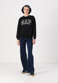 Juodas megztinis su balta "GAP" logo, derinamas su tamsiomis plačiomis džinsomis ir juodais batais. Aprangą užbaigia kibiro skrybelė.