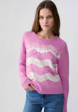 Kvinde iført en lyserød sweater med hvide paliet zigzag-mønstre og blå jeans, stående med den ene hånd i lommen på en ensfarvet baggrund.