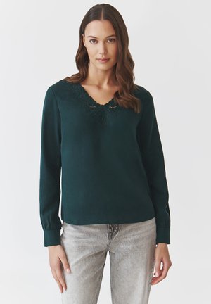 Dark green long-sleeve blouse with a V-neck, featuring floral embroidery around the neckline. Smooth texture, relaxed fit, paired with light gray jeans.