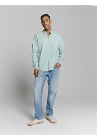 Jonge man staat ontspannen in een lichtgroen shirt met mandarin collar, lichte spijkerbroek en witte sneakers voor een effen grijze achtergrond.