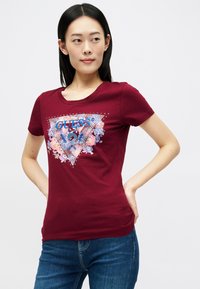 Μαρουλί μπλούζα από βαμβάκι με τριγωνικό σχέδιο που διαθέτει floral μοτίβα, μπλε λογότυπο "GUESS" και μεταλλικές λεπτομέρειες. Κοντά μανίκια, στρογγυλή λαιμόκοψη.