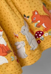 Gelber Stoff mit Punkten zeigt gestickte Tiere, darunter einen grauen Hasen und einen orangefarbenen Fuchs, sowie Blumen und Pilze.