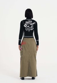 Schwarzer Strickpullover mit grafischem Design auf dem Rücken, kombiniert mit einem langen olivgrünen Cargo-Rock mit Stickerei und Seitentaschen.