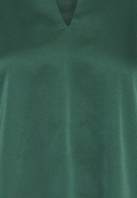 Top verde a scollo a V realizzato in un tessuto liscio e testurizzato, caratterizzato da una leggera lucentezza e un design semplice ed elegante, privo di dettagli aggiuntivi.