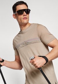 Beige t-shirt met een bergafbeelding in zwart en gekleurde strepen. Bril met donkere glazen. Handen die hikingstokken vasthouden met zwarte banden.