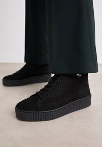 Zapatillas altas de ante negro con suelas de plataforma texturizadas, que presentan cordones redondos, detalles de costura sutiles y un pequeño acento de logo.