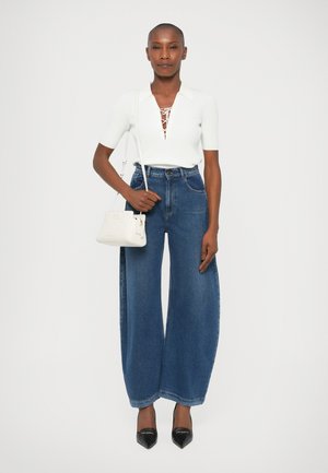 Top alb cu maneci scurte și textură canelată, cu detalii cu șiret, asortat cu blugi blue denim cu picior larg. Modelul ține o geantă mică albă și poartă tocuri negre ascuțite.