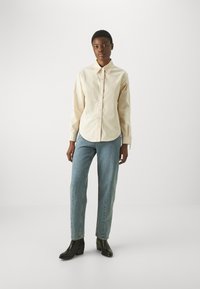 Lichtbeige button-up shirt met een klassieke kraag, voorzien van puntige schouderdetails, gecombineerd met lichtblauwe rechtgesneden jeans en zwarte enkellaarsjes met hak.