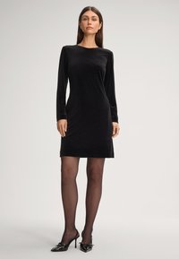 Schwarzes Samtkleid mit langen Ärmeln, einem runden Ausschnitt und gerader Silhouette, kombiniert mit schwarzen High-Heels.