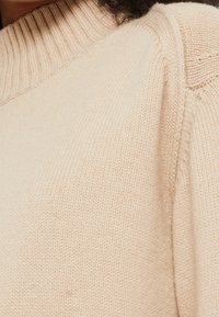 Pull en tricot beige avec un col côtelé et un motif texturé. Le tissu semble doux et est cousu avec des coutures détaillées sur les épaules.