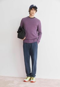 Paarse gebreide trui met structuur, marineblauwe broek met trekkoord, gele en oranje sneakers, met een zwarte tas in de hand; casual outfit tegen een effen achtergrond.